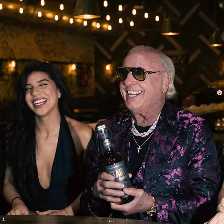 A smiling woman in a black halter top sits beside Ric Flair wearing sunglasses and a purple-and-black patterned blazer, who is holding a bottle labeled 'LAIR'. The photo is taken in a dimly lit bar or lounge with warm string lights and wood-toned decor in the background.