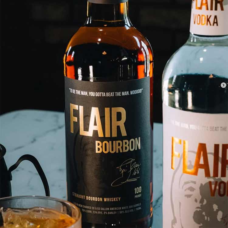 A bottle of FLAIR Bourbon Straight Bourbon Whiskey in the 100 Proof variant, shown next to another bottle of FLAIR Vodka on a light marble surface with a glass of whiskey in the foreground.