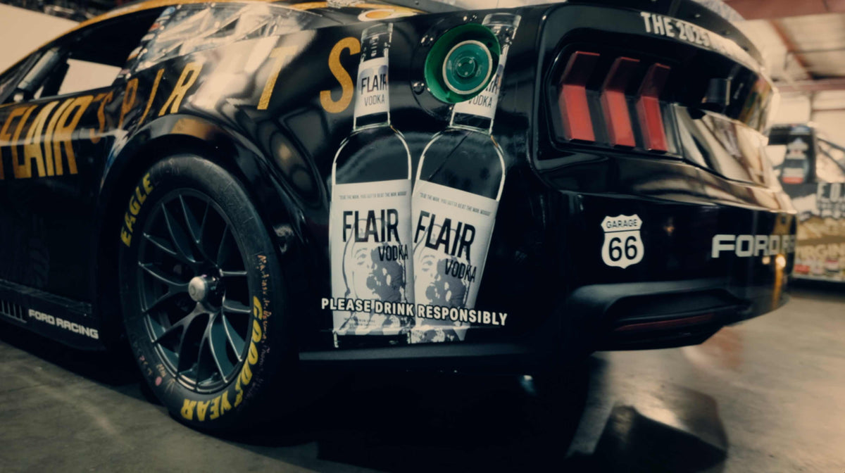A black Ford race car in a garage with two large bottles of Flair Vodka hanging on the rear bumper. The car features gold and white racing graphics, including 'THE ZONE', 'FORD', and 'RACING'.
