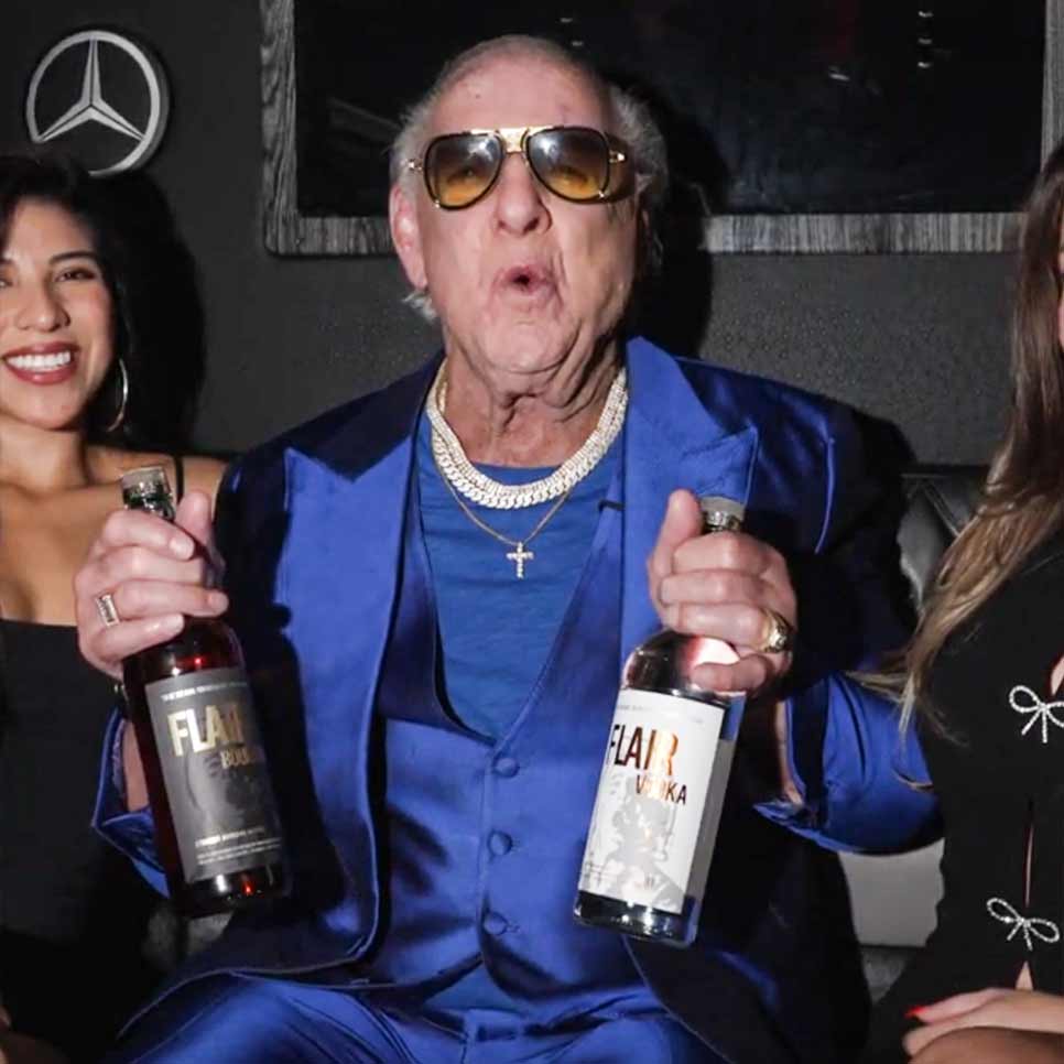 Ric Flair in a shiny blue suit and sunglasses holds two bottles labeled 'FLAIR Vodka' in a dim indoor setting, with two women partially visible at the sides.