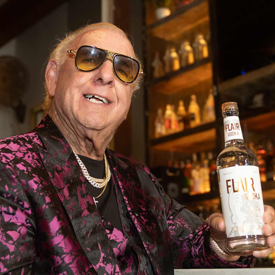 World Champion Ric Flair wearing gold-framed sunglasses, a black shirt, and a pink-and-black patterned blazer holds a bottle of FLIR VODKA in a bar setting with shelves of bottles in the background.